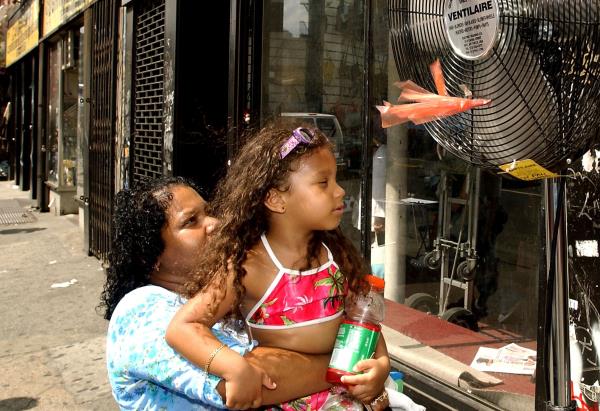 A woman holds her young daughter up to the fan on a street with storefro<em></em>nts and lots of pavement.