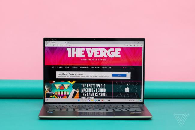 The Acer Swift 5 open on a green and pink background. The screen displays The Verge homepage.