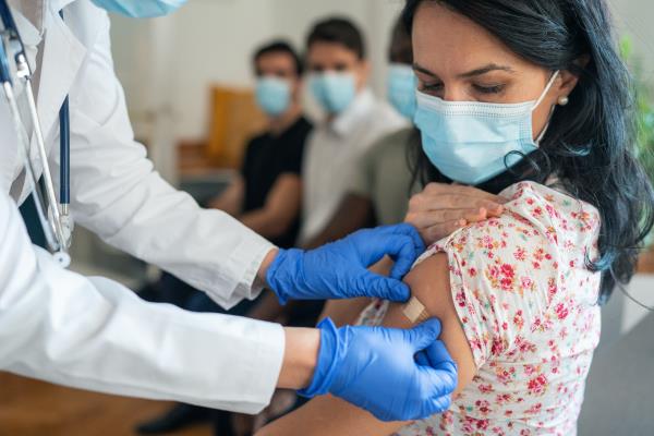 A doctor in white lab coat and blue gloves places Band-Aid on a woman wearing a mask who has just received a COVID-19 vaccine.
