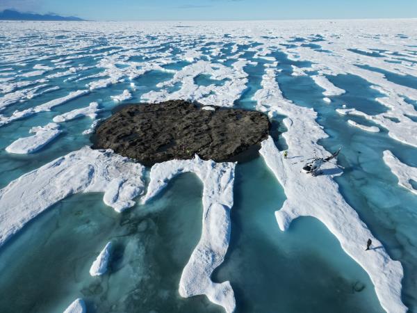 A helicopter sits on ice next to what appears to be a gravel-covered island, but is actually an iceberg. People are standing on its surface.